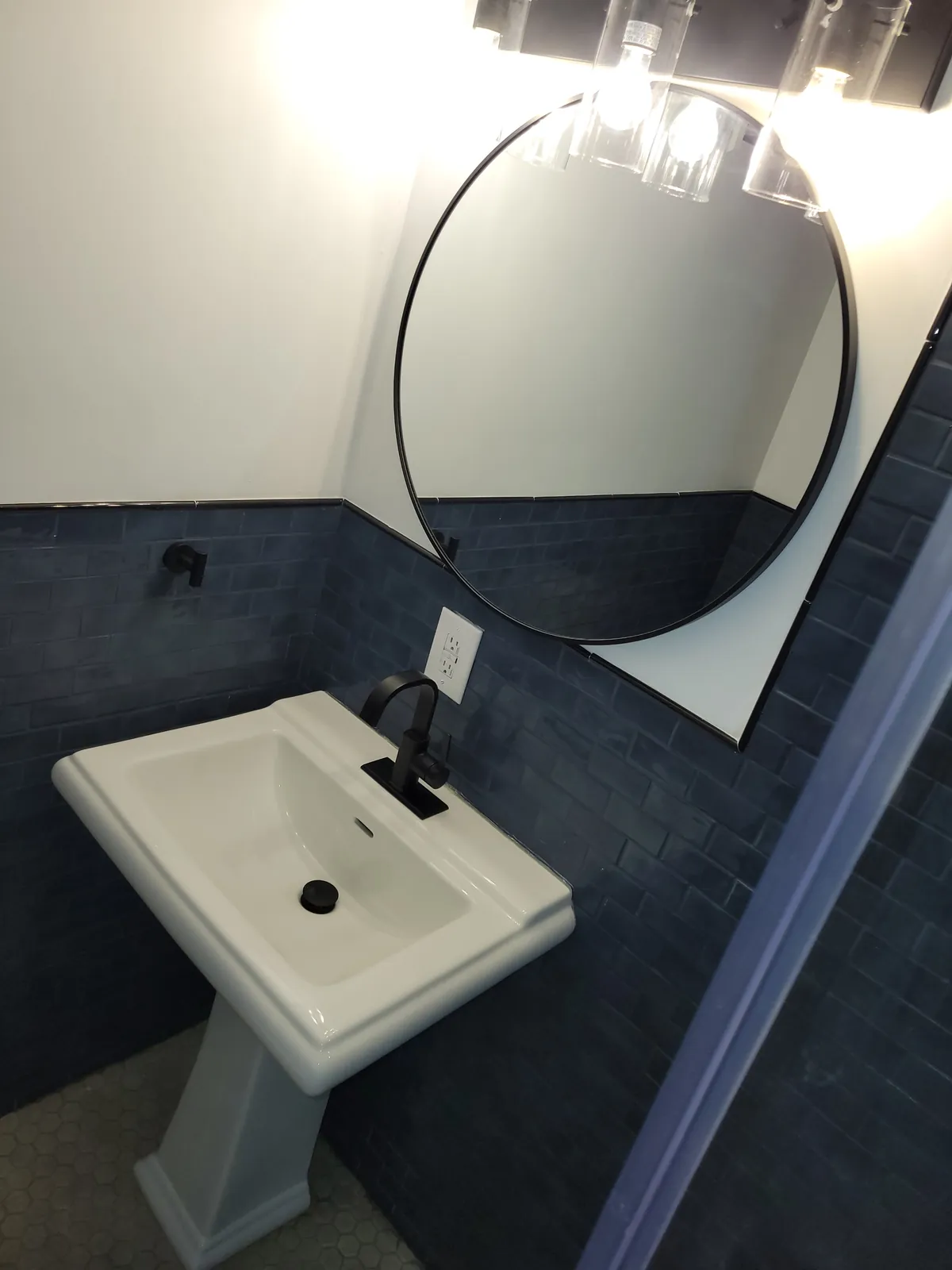 Bathroom pedestal sink and round black mirror against a dark blue tile wall
